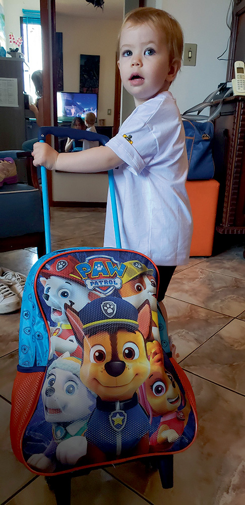 Uma bebê aparece em pé segurando uma mochila de rodinha temática da Patrulha Canina. Ela usa uma camiseta branca e está na sala de casa.