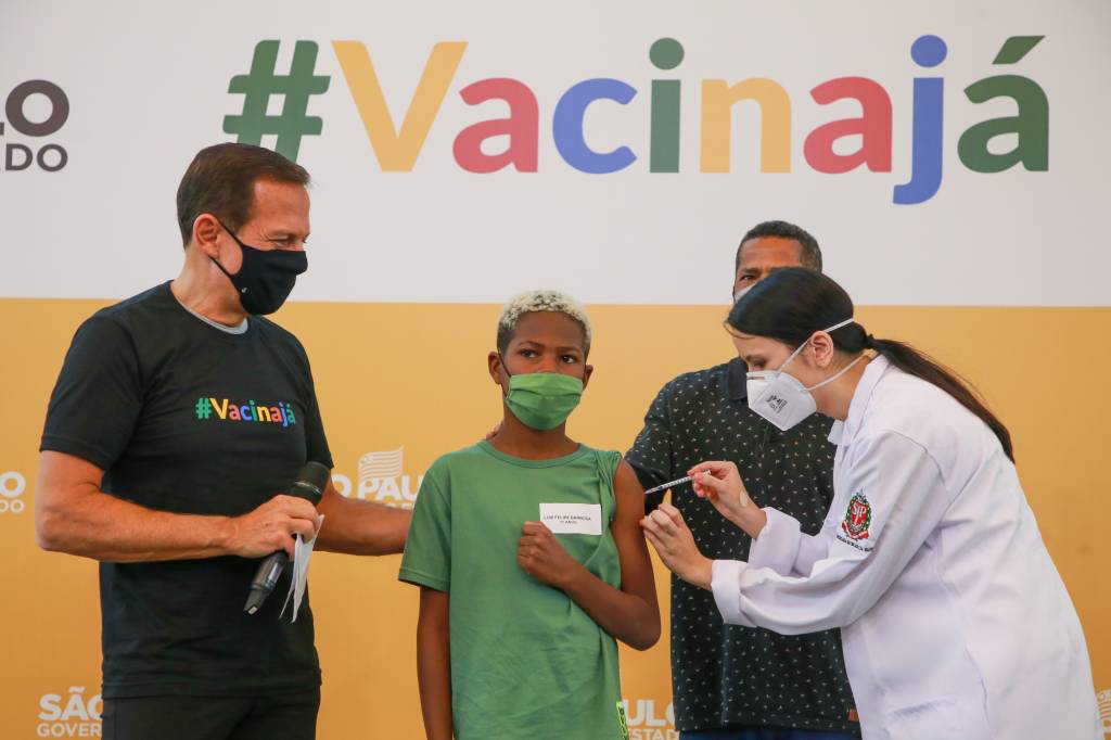 Imagem mostra garoto de camiseta verde recebendo vacina no braço. Uma enfermeira aplica a dose, enquanto dois homens ficam de pé ao lado do menino. Ao fundo, um banner escrito "Vacina Já"