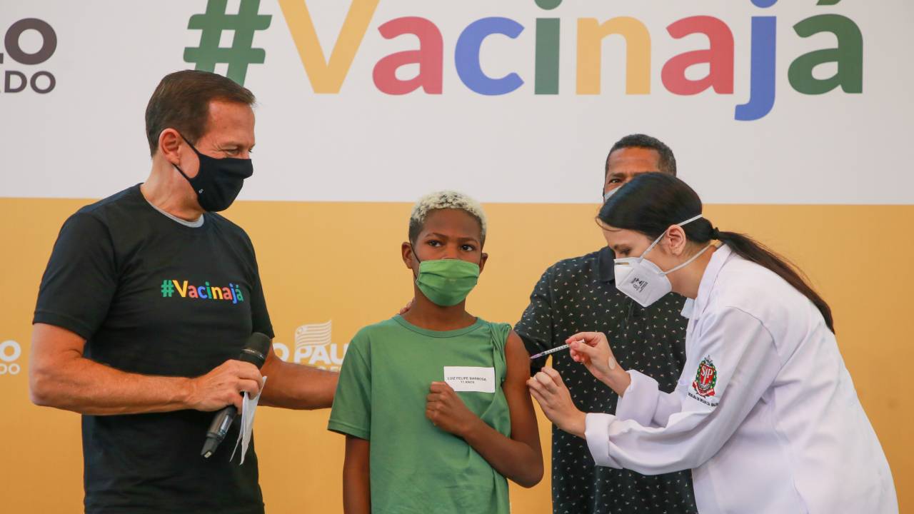 Imagem mostra garoto de camiseta verde recebendo vacina no braço. Uma enfermeira aplica a dose, enquanto dois homens ficam de pé ao lado do menino. Ao fundo, um banner escrito "Vacina Já"