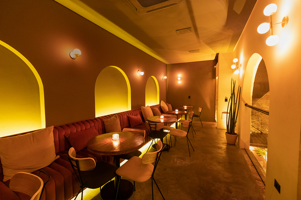 Ambiente de bar levemente iluminado por velas nas mesas posicionadas em frente a um estofado de cor vermelha na parede