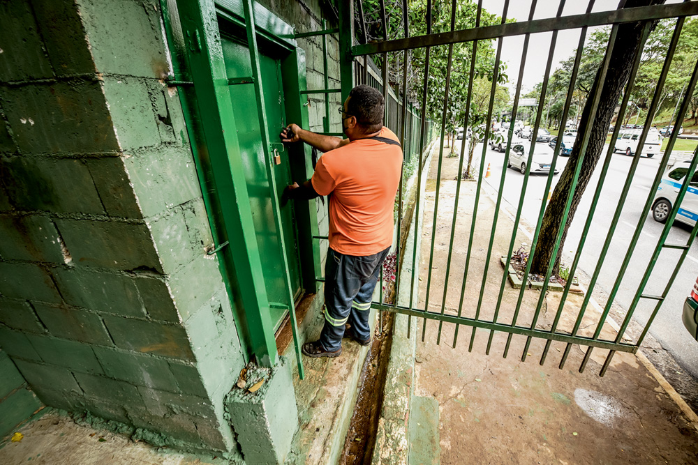 Imagem mostra homem de camiseta laranja trancando porta verde com grades de segurança.