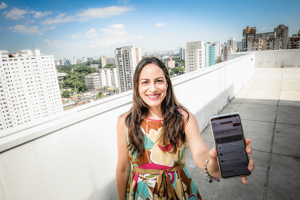 Catarina Anderáos aparece mostrando seu telefone celular para a câmera, em uma sacada de um condomínio. Ela é uma mulher branca, de cabelos e olhos castanhos e usa um vestido colorido e estampado.