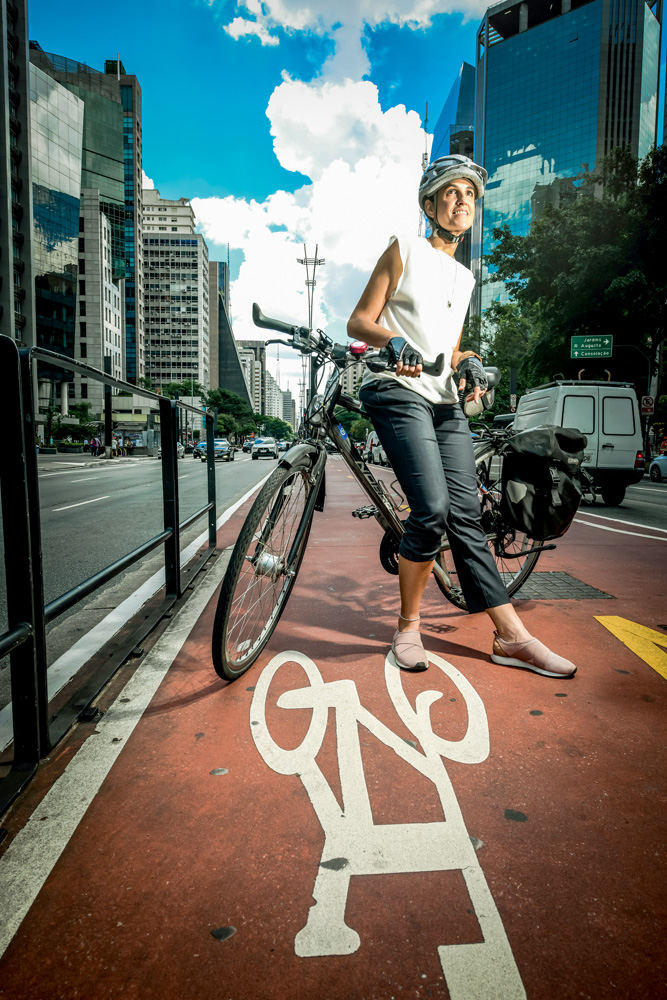 Imagem mostra mulher de camiseta branca e calça preta apoiada sobre sua bicicleta, em uma ciclovia entre duas faixas de carro.