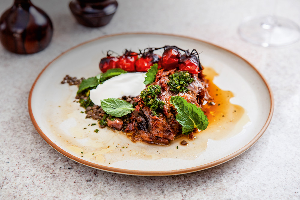 Cordeiro servido junto de tomates assados, folhas de hortelã, lentilha e coalhada em um prato raso de louça branca com borda em tom de marrom