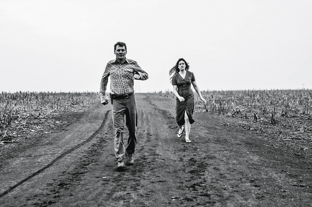 Foto em preto e branco mostra um homem e uma mulher andando em uma estrada vazia. Não há nada em volta, só uma vegetação rasa no chão. Ambos são brancos. Ela anda atrás dele.