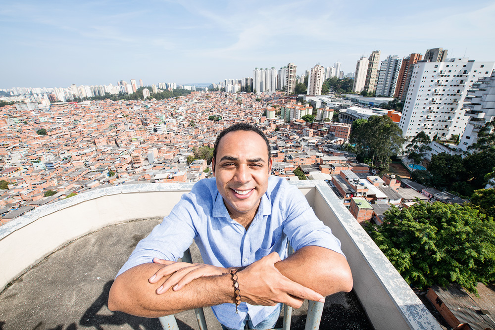 Imagem mostra homem de camisa azul e os braços cruzados. Ao fundo, uma favela.