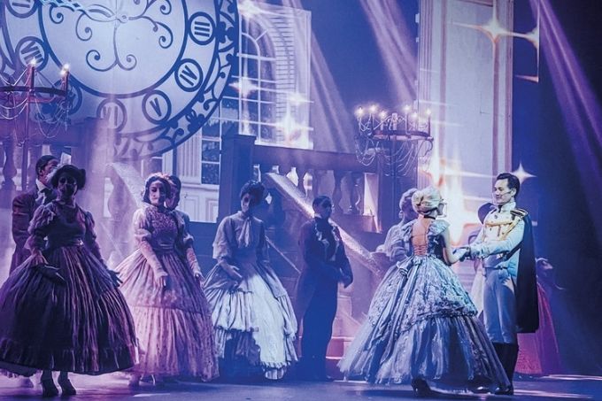 Foto mostra Cinderella e o príncipe no teatro. Eles contracenam com outros membros do baile. Uma luz azul intensa cobre o palco.
