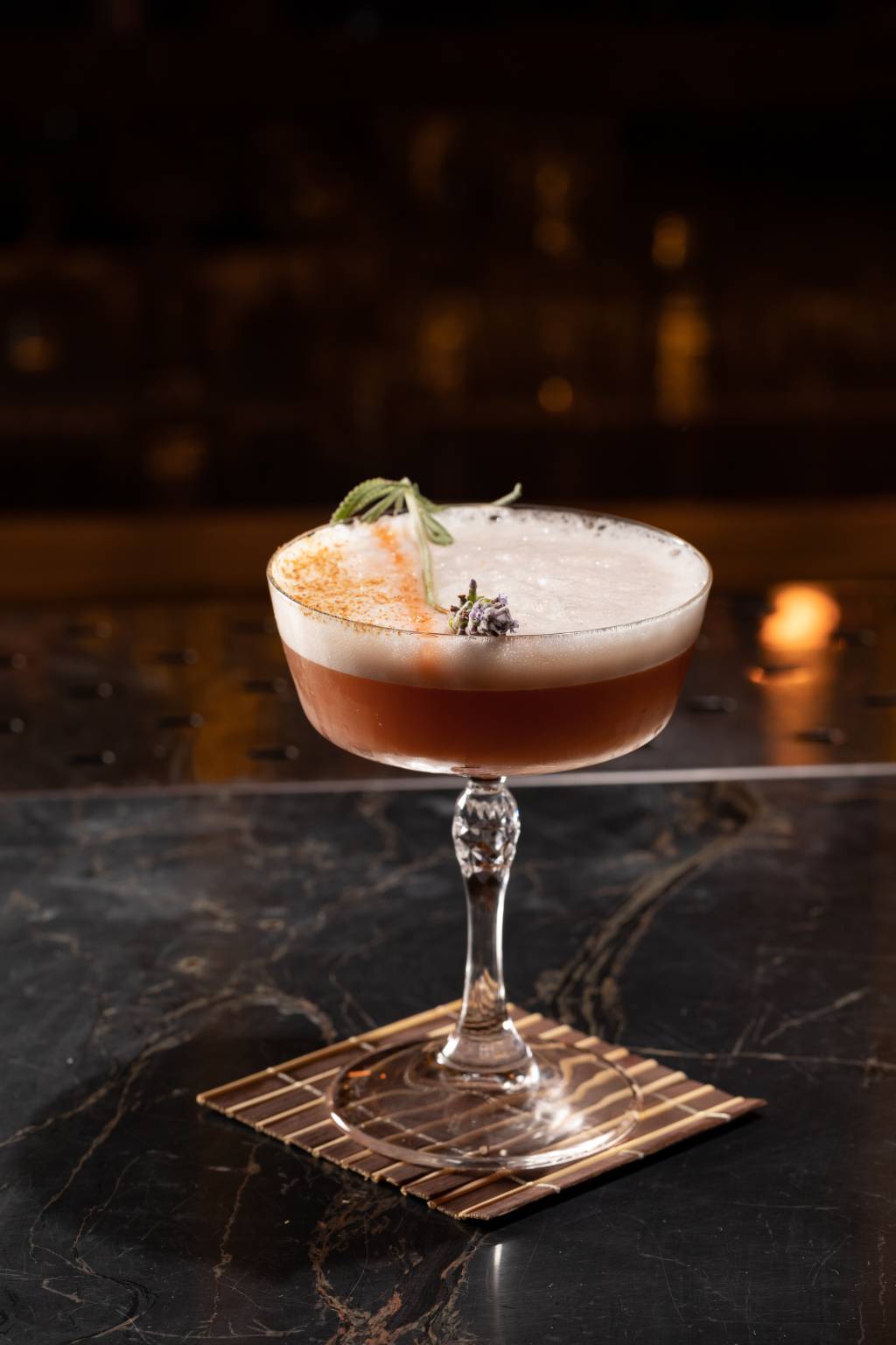 Carrasco Drinque de cor levemente alaranjada com uma camada de espuma no topo e decorado por uma pequena flor de cor lilás