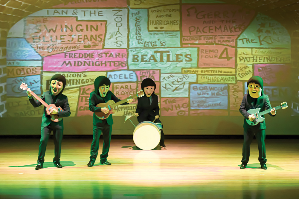 Foto mostra 4 pessoas com fantasias dos Beatles ao estilo cartoon em cena. Eles seguram instrumentos e se apresentam em um palco com luzes coloridas.