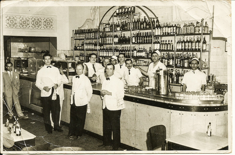 Imagem mostra balcão de bar com diversos garçons de camisas brancas, calças prestas e gravatas borboletas.