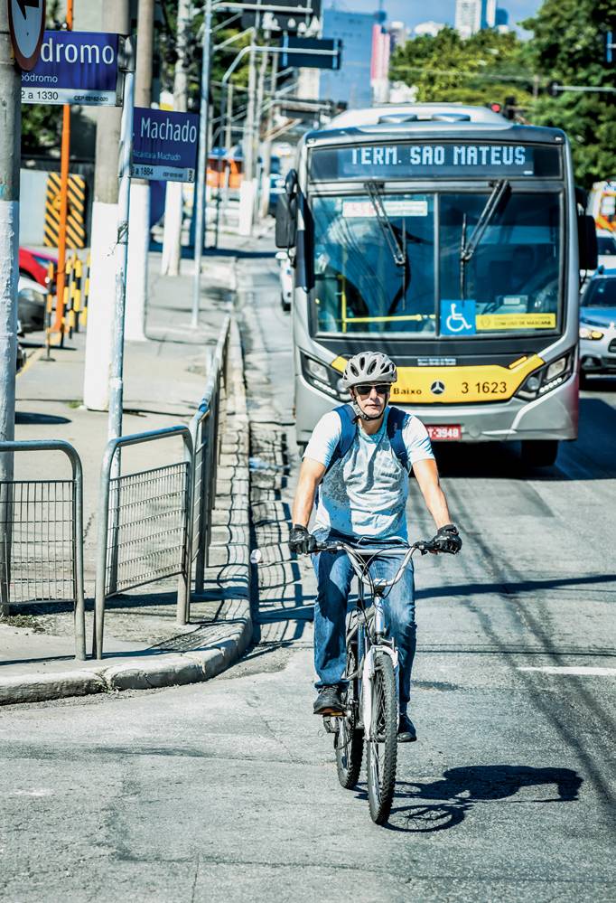 Imagem mostra homem em bicicleta, de capacete e camiseta azul, na rua. Atrás dele, um ônibus amarelo.