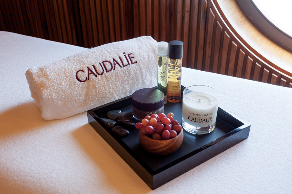 Imagem mostra bandeja com cosméticos, uma vela e um cacho de uvas, sobre cama. Uma toalha enrolada também está sobre a cama.