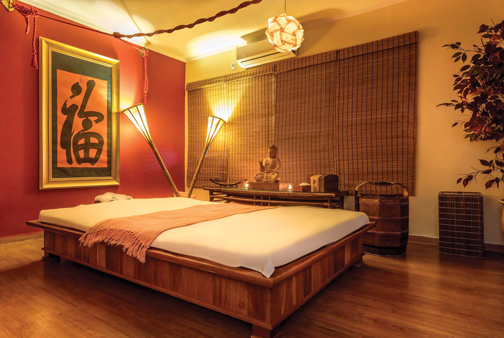 Buddha Spa – Jardins Imagem mostra quarto iluminado por três luminárias amarelas. Uma das paredes, ao fundo, é vermelha e tem um quadro. No centro do quarto, uma cama com estrutura de madeira, assim como o piso.