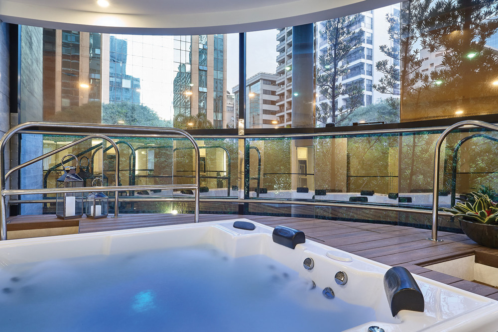 FOTO 7 – The Spa at Renaissance – FOTO DIVULGACAO.jpg Imagem mostra, na parte inferior, jacuzzi com luz azul sob a água. No resto da imagem, uma janela grande de vidro que mostra os outros prédios ao redor.