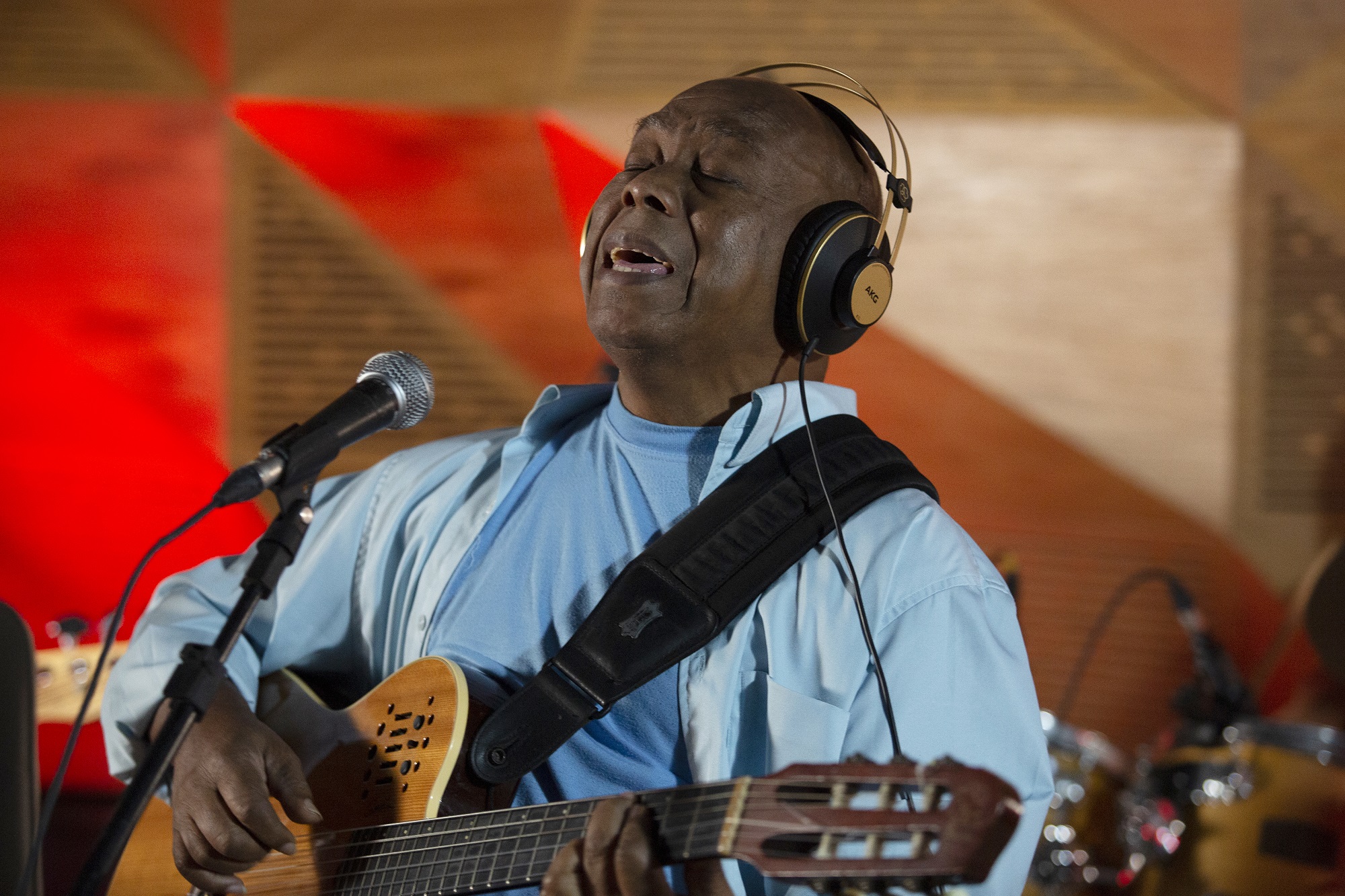 Imagem mostra homem de camisa e camiseta azul segurando um violão e cantando em um microfone. Ele também usa um fone.