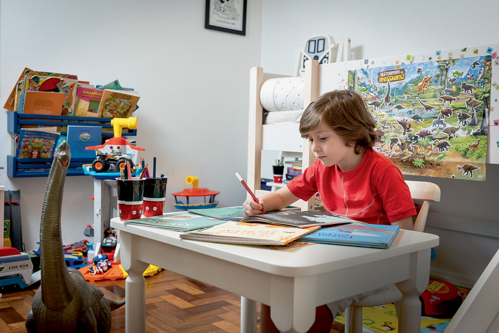 Gabriel aparece sentado em uma mesa branca. Ele escreve em um caderno. O ambiente ao redor é decorado com brinquedos.