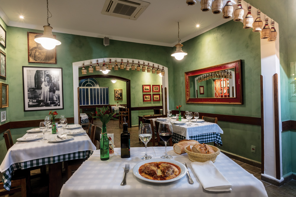 Imagem mostra salão com paredes verdes e mesas com pratos de macarrão servidos.