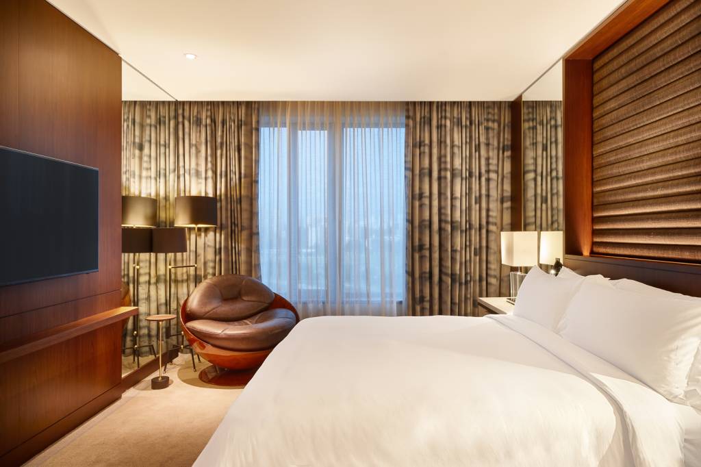 Foto de quarto de hotel com cama grande de casal à direita. Poltrona de couro marrom no canto esquerdo ao fundo.