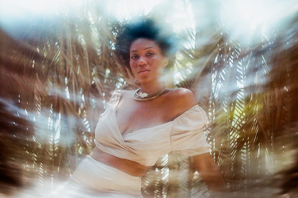 Imagem msotar mulher de vestido branco, em frente à folhas de palmeiras.
