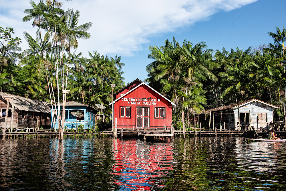 Foto de Luiz Braga mostra uma casa a beira de um rio na Amazônia. A casa é vermelha e está cercada por outras.