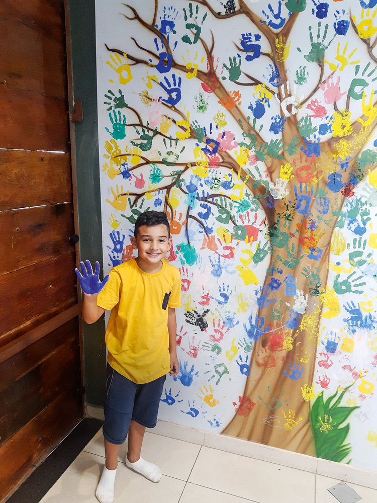 Foto de Miguel, que veste uma camiseta amarela e um shorts azul, em frente a um mural colorido pintado com a forma de suas mãos.