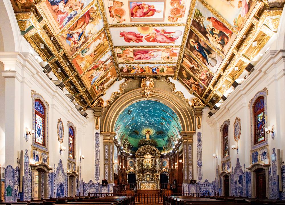 Imagem mostra salão de igreja com paredes laterais brancas e azuis e teto com afrescos de cenas bíblicas. Ao fundo, o altar da Igreja, com um teto azulado.