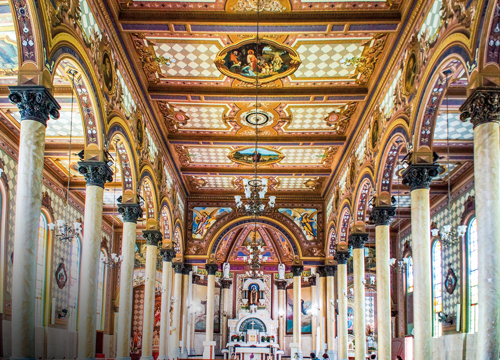 Imagem mostra salão de igreja com teto marrom e branco, com imagens bíblicas entre padrões quadriculados. Duas fileiras de colunas de mármore acompanham o salão até o fundo, onde está um altar.