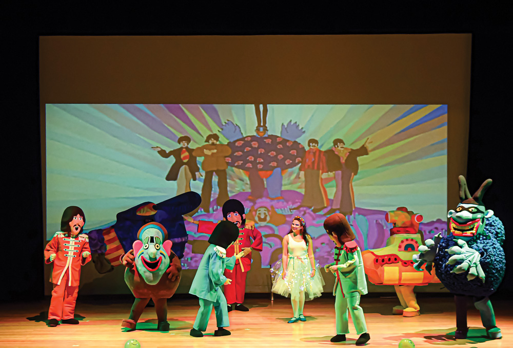 Foto mostra 4 pessoas com fantasias dos Beatles ao estilo cartoon em cena. Eles se apresentam em um palco com luzes coloridas. Outros personagens do filme 