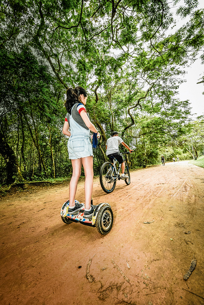 Imagem mostra garota em patinete e menino em bicicleta andando em estrada de terra.