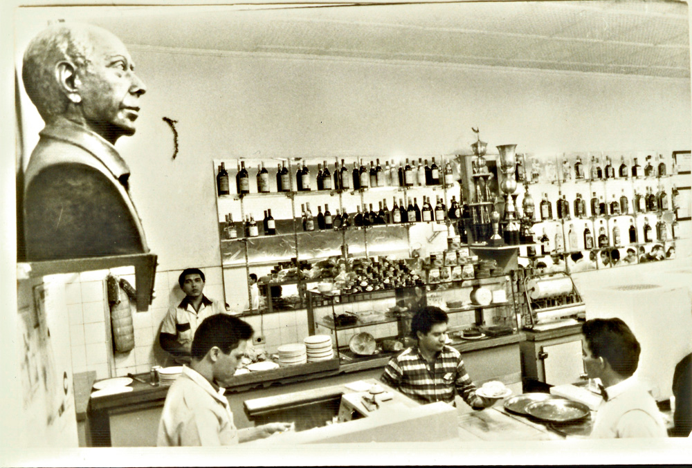Imagem em preto e branco mostra bar com diversos funcionários e um busto metálico exposto na parede.