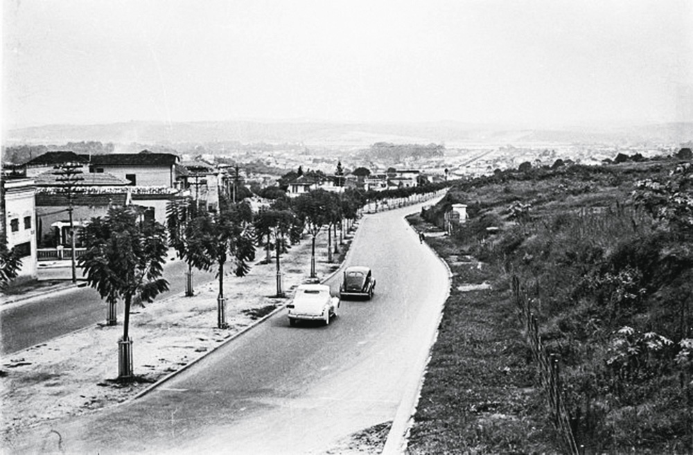 Imagem em preto e branco mostra rua, ao centro, com dois carros trafegando. À direita da rua, mato, à esquerda, casas.