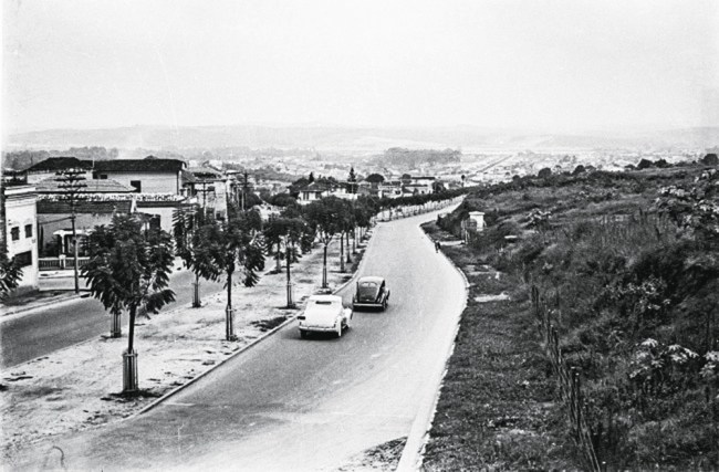 Imagem em preto e branco mostra rua, ao centro, com dois carros trafegando. À direita da rua, mato, à esquerda, casas.