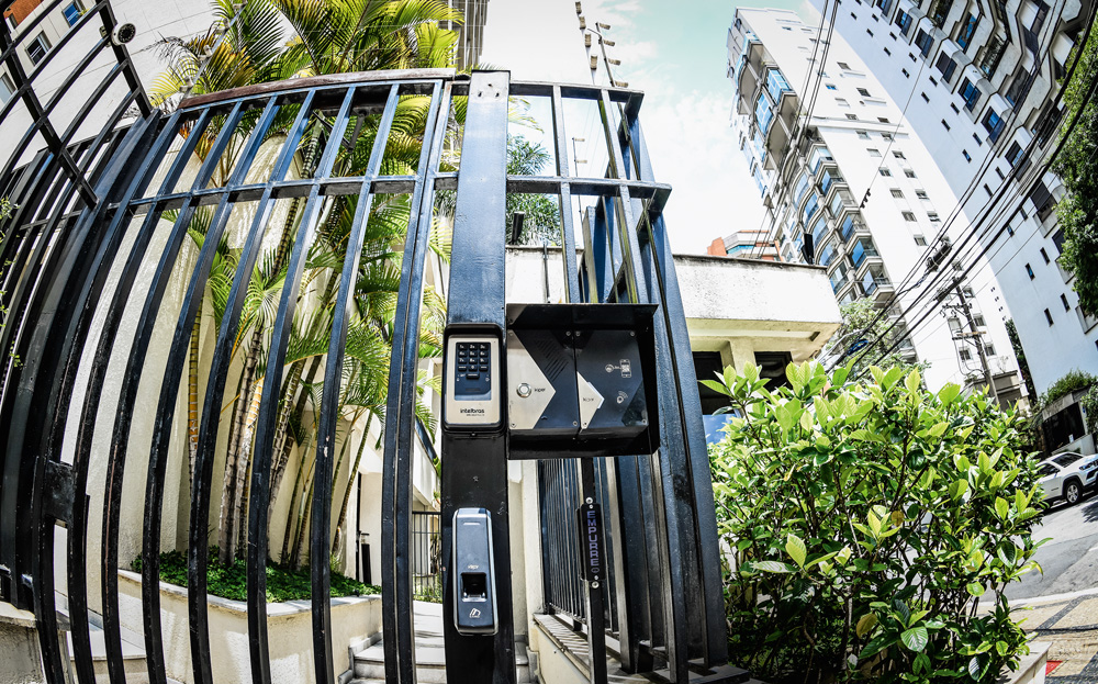 Foto mostra um portão de grades pretas de uma portaria de condomínio. No portão, um dispositivo de reconhecimento facial ministra a entrada e saída de pessoas.