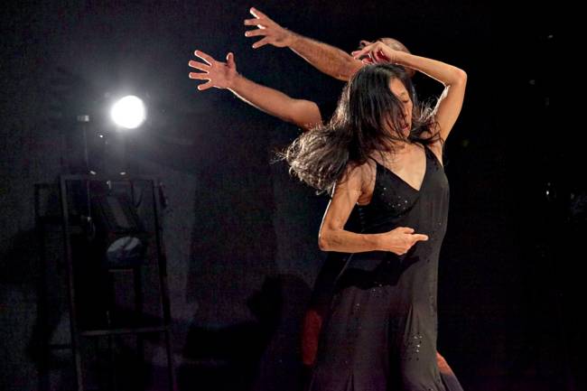 Imagem mostra mulher de vestido preto dançando . Ao fundo, um foco de luz branco.