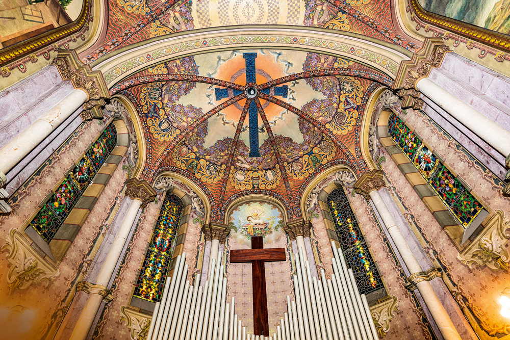 Imagem mostra teto de igreja com uma cruz azulada ao centro e pinturas geométricas ao seu redor, coloridas. Nas laterais, vitrais também coloridos e, na parte inferior da imagem, uma cruz de madeira e alguns tubos de um orgão.