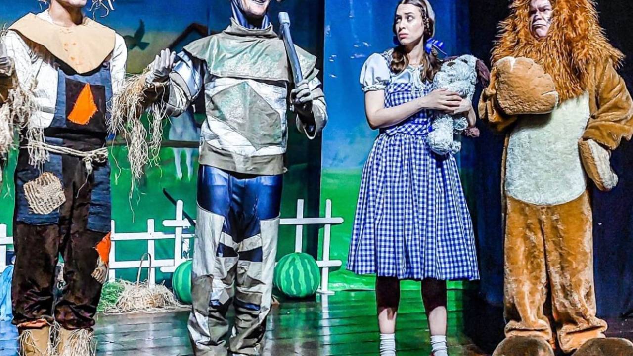 Foto dos atores de "O Mágico de Oz" em cena.Dorothy, de vestido azul e sapatos de boneca, está ao lado do Homem de Lata, Espantalho e Leão.