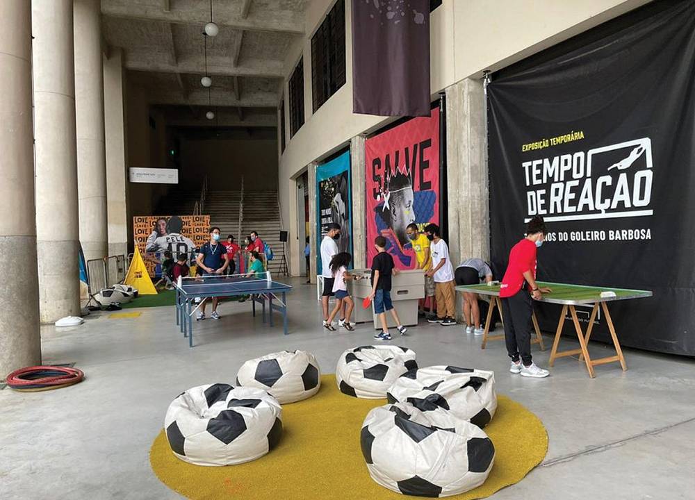 Foto mostra a área externa do museu, com vários puffs em forma de bola de futebol, mesas de pebolim e outras atividades. Crianças e adultos de máscara brincam no espaço.