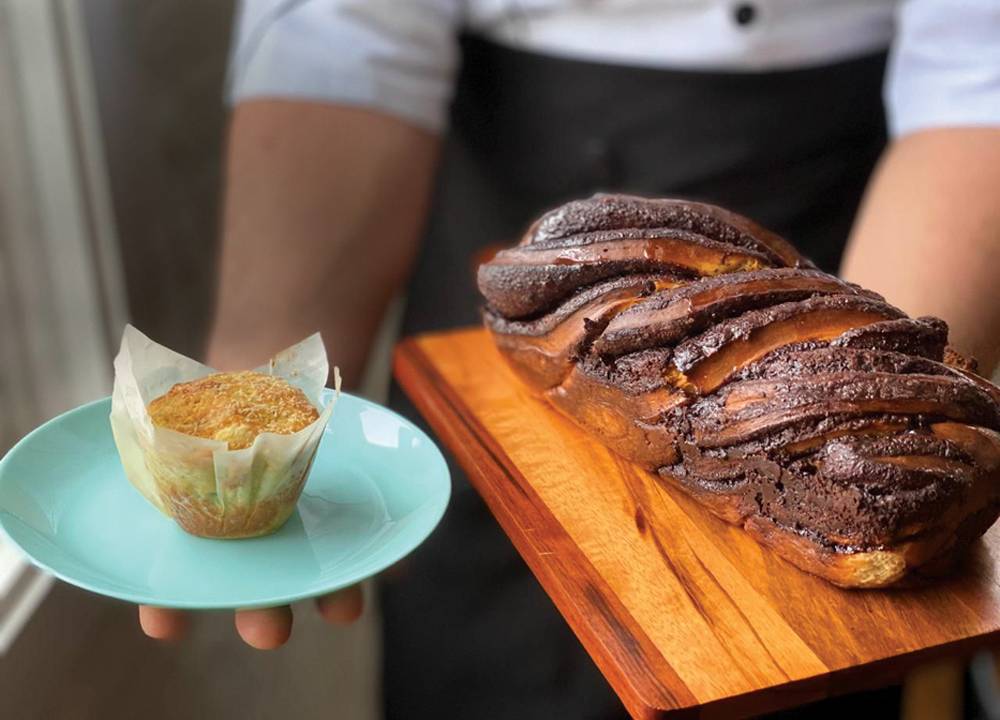 Lado a lado, muffin servido em prato raso de louça azul e babka de chocolate, pão doce trançado, servido sobre tábua de madeira