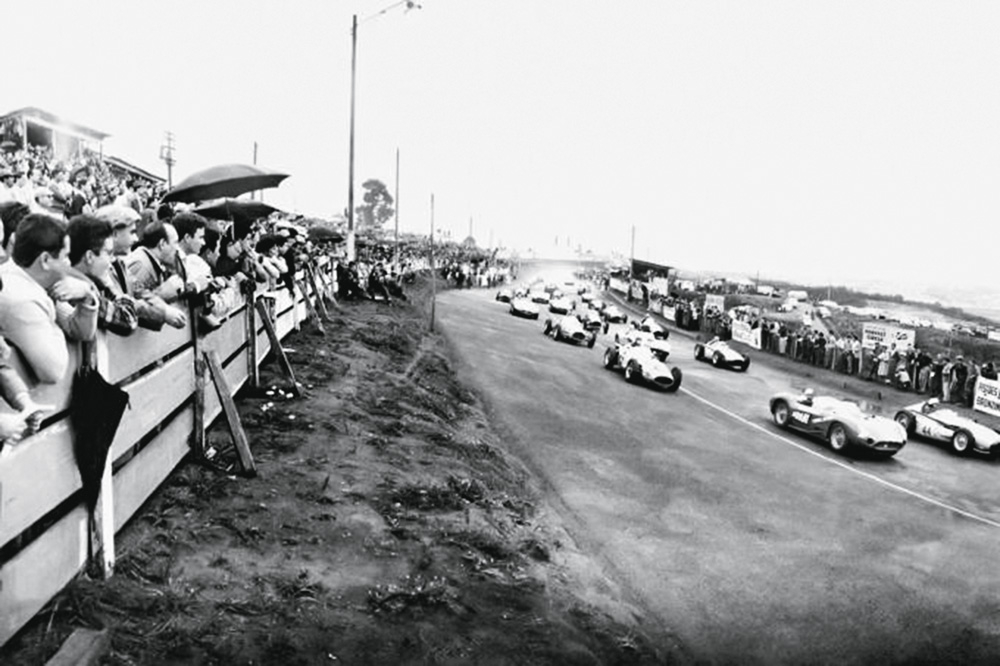 Imagem mostra carros de corrida em pista, à direita, e multidão atrás de barreira de madeira, à esquerda,