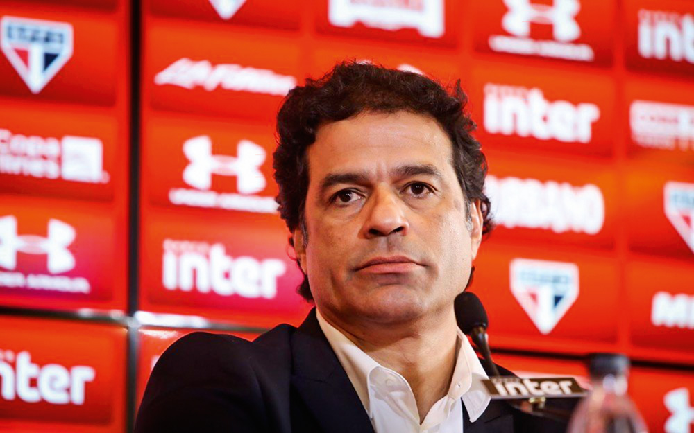 Imagem mostra homem de terno em frente a microfone, com um fundo vermelho cheio de logos de marcas e do clube São Paulo.