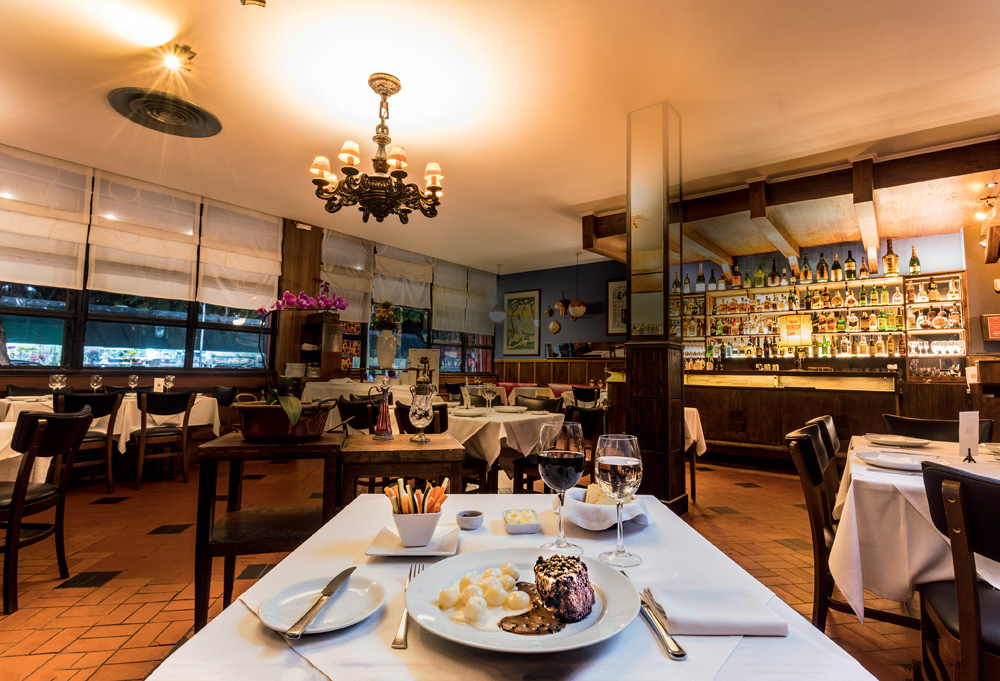 Imagem mostra espaço com mesas e cadeiras, uma parede azul ao fundo e janelas. À frente na foto está um prato com carne e batatas e duas taças, uma de vinho e outra com água.