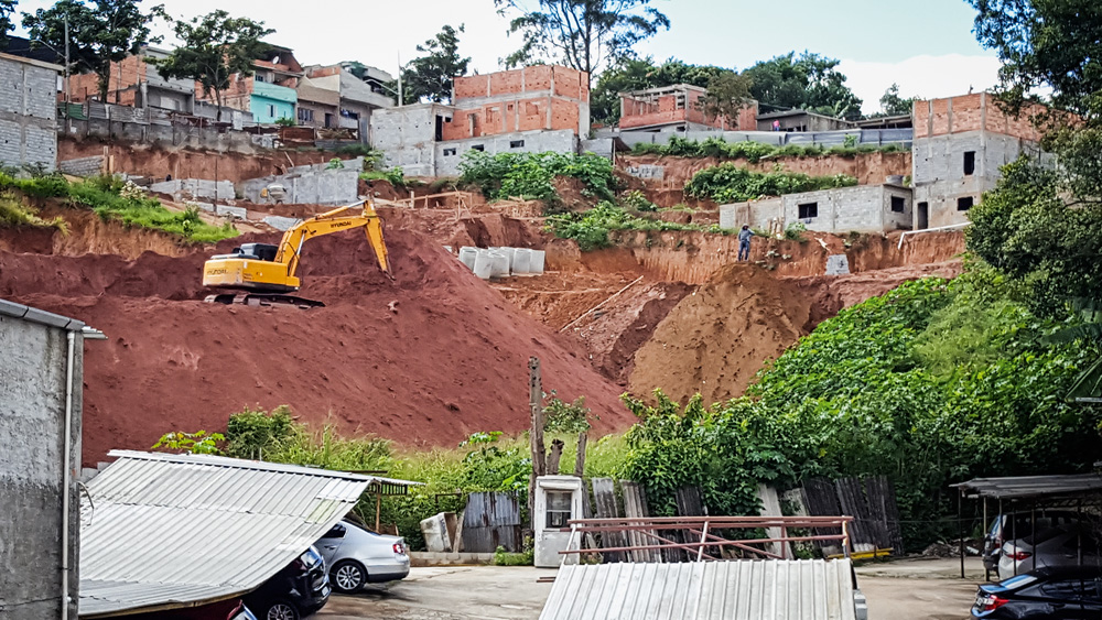 Foto mostra uma escavação em um terreno irregular na Zona Sul de SP. Uma escavadeira amarela faz o serviço. Em volta do morro, há casas e árvores.