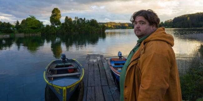 Um homem olha para a câmera; atrás dele, barcos e um lago