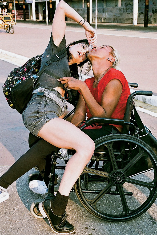 Foto de uma performance mostra uma mulher sentada em cima de um homem em uma cadeira de rodas.
