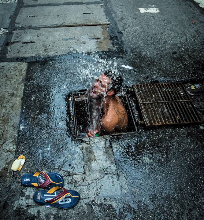 Foto mostra um morador de rua usando água de um bueiro para tomar banho.