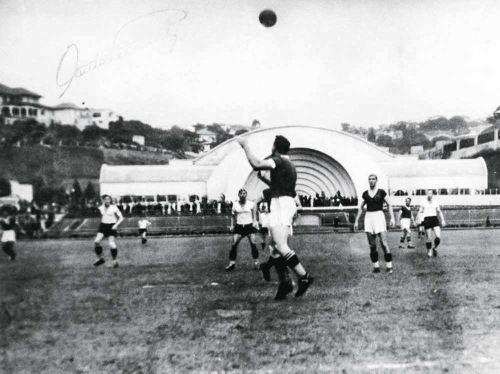 Imagem mostra jogadores de futebol em gramado. Ao fundo, uma estrutura branca.