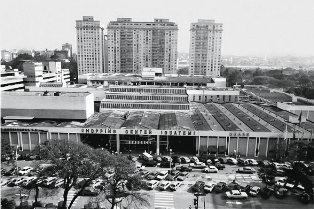 Imagem mostra espaço enorme com carros estacionados à frente. Ao fundo, prédios.