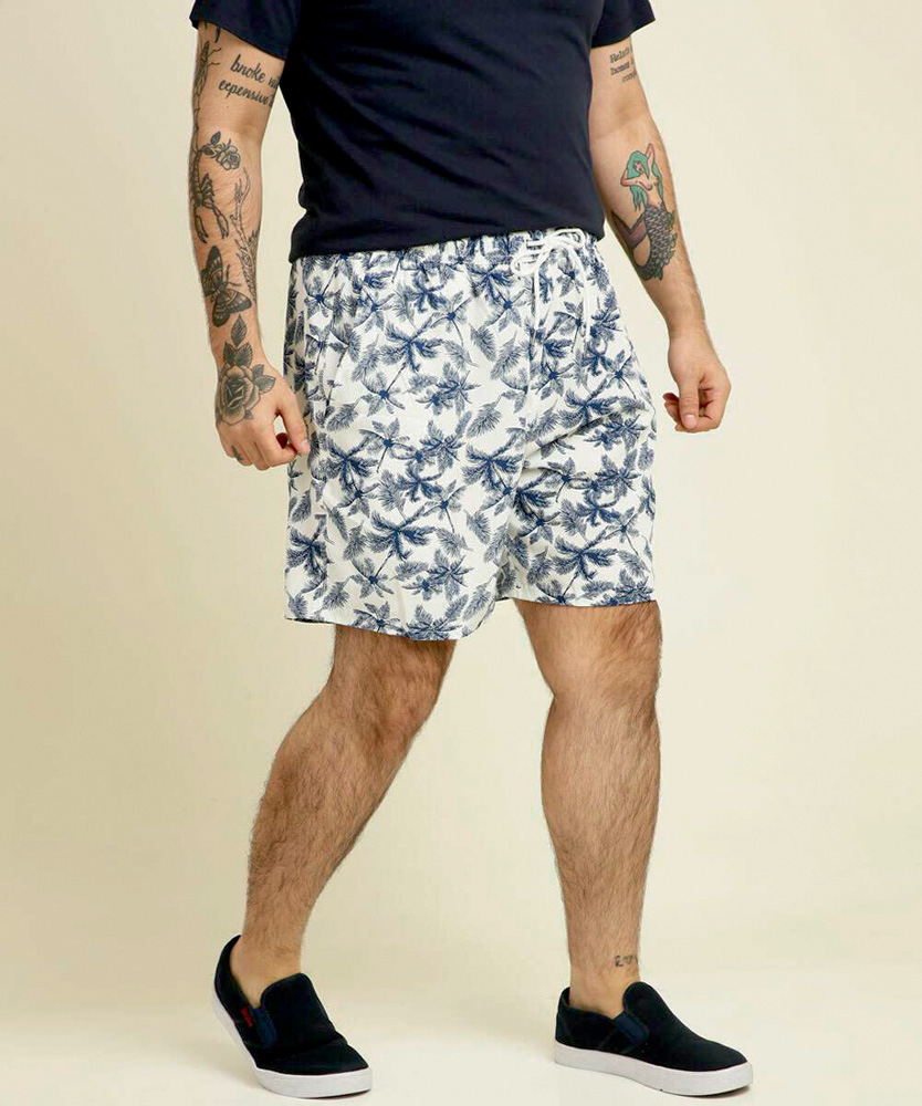 Modelo aparece do ombro para baixo usando uma bermuda floral em tons de azul. Ele é branco e tem tatuagens no braço.