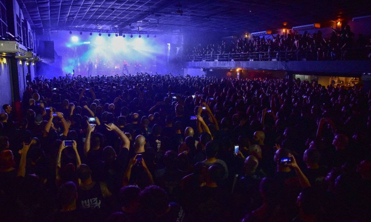 Imagem mostra espaço com multidão de pessoas e palco iluminado de cores azuis ao fundo.