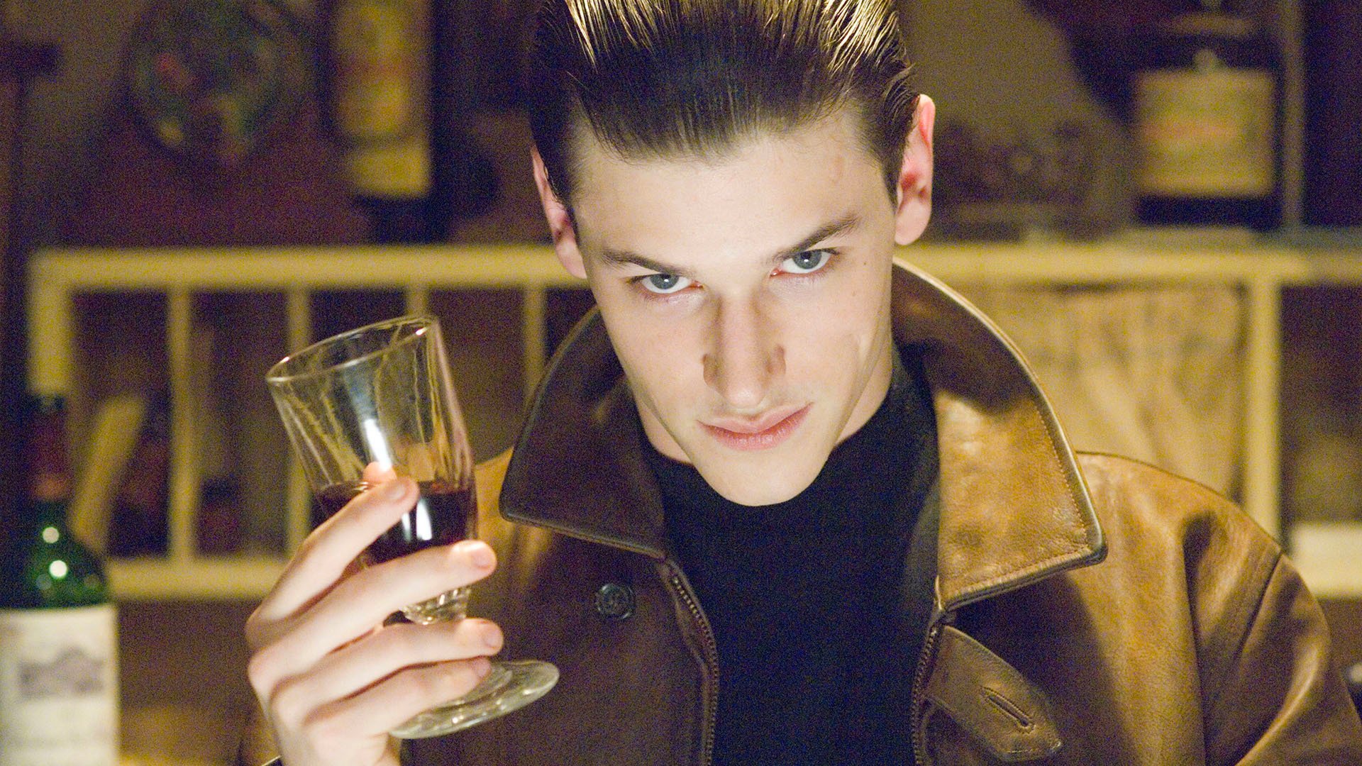 Imagem mostra um homem de jaqueta marrom, segurando um copo de vinho e olhando para a câmera.